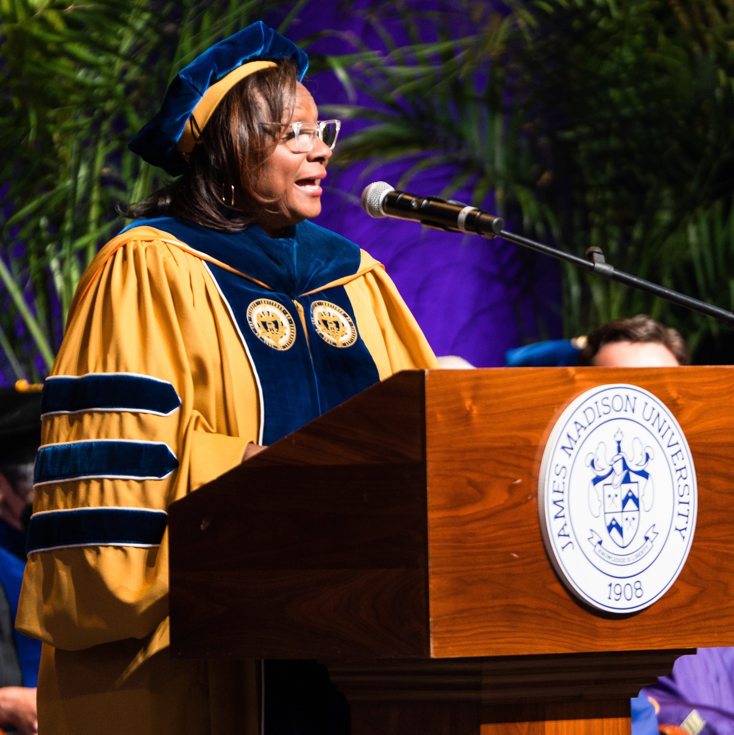Dr. Linda M. Thomas speaks into a microphone at the podium for commencement at James Madison University. She is wearing doctoral regalia.