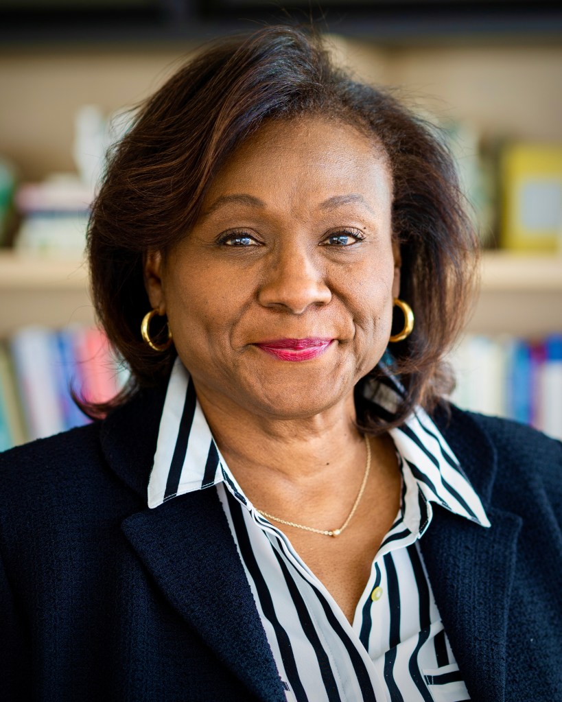 A headshot of Dr. Linda M. Thomas, an African-American Professor and University Dean, smiling in front of a bookcase. Linda is wearing a blazer and a white and black striped button-down with a thin chain necklace and small gold hoop earrings.